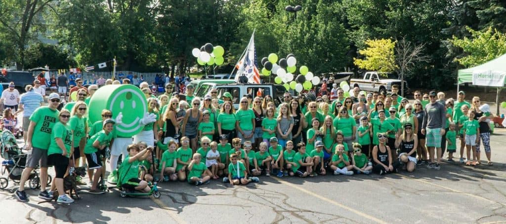 Happy outdoor community event with families and volunteers, promoting local real estate and housing in Michigan, featuring a large group photo with balloons, banners, and festive atmosphere.